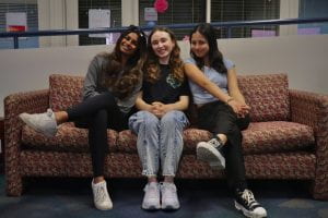 Three young women sitting next to each other on a red couch. Starting from the left side, girl 1 with dark brown hair, wearing a gray pullover sweater, black leggings, and white sneakers. Girl 2 in the middle has reddish brown hair and is wearing a black shirt, jeans, and white sneakers. Girl 3 on the right is wearing a light blue shirt, black pants, and black and white sneakers. All girls are smiling towards the camera.
